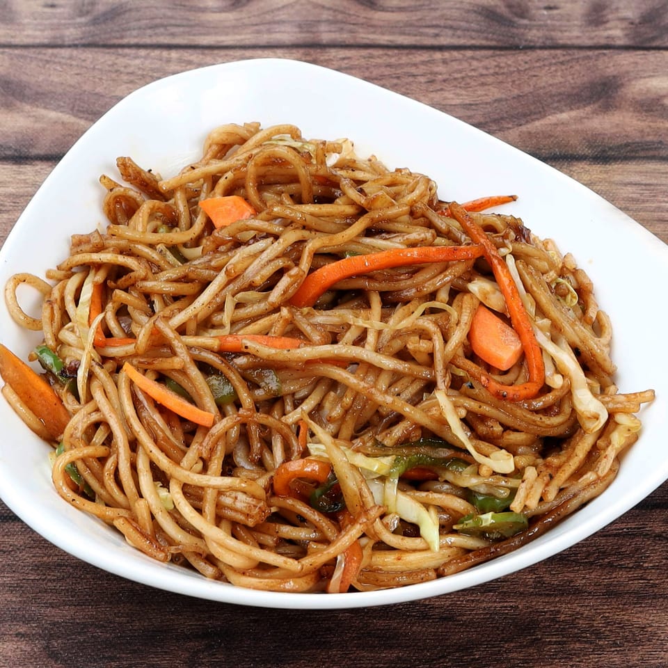 A white bowl filled with Everything Noodles, featuring stir-fried noodles, carrots, and cabbage on a wooden table.