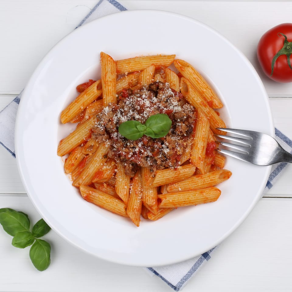 Penne pasta with Bolognese sauce, grated cheese, and basil on a white plate with a fork.